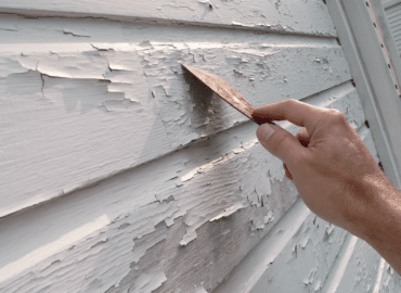 a hand removing peeling paint from the exterior of a home with a scraper tool.