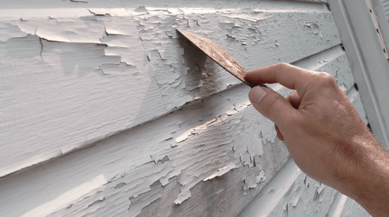 a hand removing peeling paint from the exterior of a home with a scraper tool.